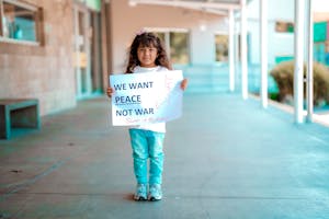 Little girl with long hair standing on pavement holding a 'We Want Peace Not War' sign.